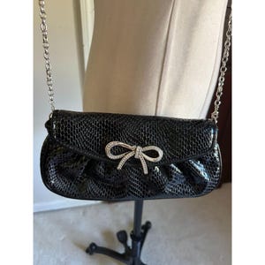 Puede incluir: Bolso de mano negro con una correa de cadena plateada. El bolso presenta un patr&oacute;n texturizado similar a la piel de reptil y un adorno en forma de lazo con brillantes pedrer&iacute;a. El bolso se exhibe en un maniqu&iacute;.