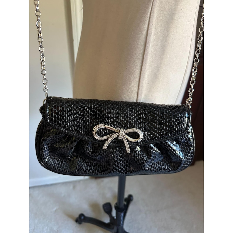 Puede incluir: Bolso de mano negro con una correa de cadena plateada. El bolso presenta un patr&oacute;n texturizado similar a la piel de reptil y un adorno en forma de lazo con brillantes pedrer&iacute;a. El bolso se exhibe en un maniqu&iacute;.