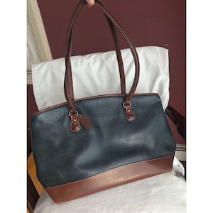 May include: A navy blue and brown leather tote bag with brown handles and trim. The bag has a rectangular shape with a rounded top and a brown leather base. A small brown leather tag hangs from the handle.