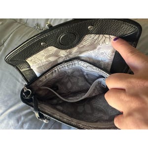 Puede incluir: Bolso de mano de cuero negro con una correa de cadena plateada. El interior presenta un forro con estampado floral en tonos grises y blancos. El bolso tiene un cierre de solapa y un peque&ntilde;o bolsillo interior. El bolso est&aacute; abierto, revelando el interior.