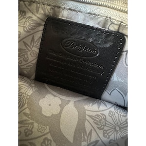 Puede incluir: Primer plano de una etiqueta de cuero negro con el nombre de la marca "Brighton" en relieve en una fuente cursiva. La etiqueta est&aacute; dentro de una bolsa con un forro estampado floral. El texto "The Brighton Collection" es visible.