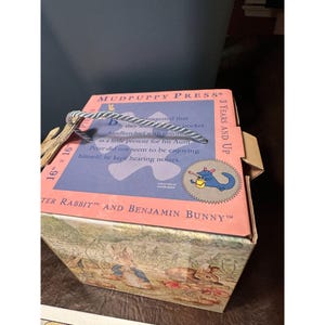 May include: A box for a 16-inch by 16-inch puzzle featuring Peter Rabbit and Benjamin Bunny. The box is pink and blue with a white rope handle. The text on the box reads "Mudpuppy Press", "16" x 16", "3 Years and Up", "Peter Rabbit" and "Benjamin Bunny".