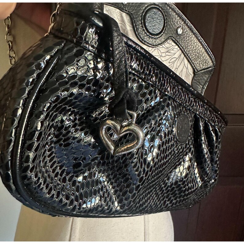 Puede incluir: Bolso de mano negro brillante con estampado de piel de serpiente. El bolso tiene un charm plateado en forma de coraz&oacute;n y una correa de cadena. El forro interior tiene un estampado. El bolso est&aacute; sobre un maniqu&iacute;.