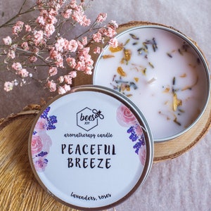 Puede incluir: Una vela de aromaterapia blanca en una lata plateada con un diseño floral. La vela está etiquetada como "Bees Joy" y "Peaceful Breeze" con el aroma "lavanda, rosas".