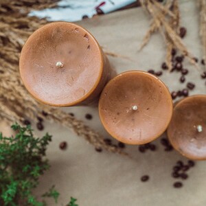 May include: Three brown pillar candles with white wicks, arranged in a row on a beige surface with scattered coffee beans.