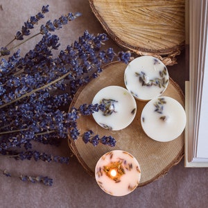 Puede incluir: Cuatro velas de té blancas con flores de lavanda secas en la parte superior, colocadas sobre una superficie de madera con un ramo de lavanda en el fondo.