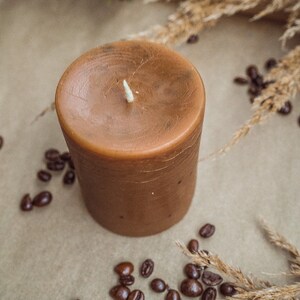 May include: A brown candle with a white wick sits on a brown surface surrounded by coffee beans and dried grass.