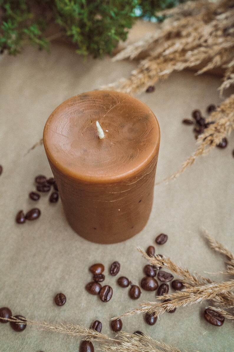 May include: A brown candle with a white wick sits on a brown surface surrounded by coffee beans and dried grass.