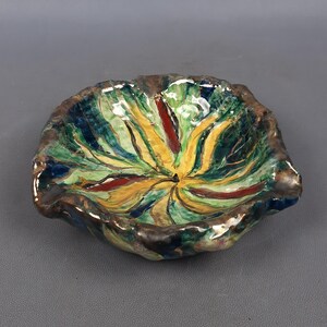 Puede incluir: Un cuenco de cer&aacute;mica pintado a mano con una forma org&aacute;nica &uacute;nica. El cuenco presenta una vibrante paleta de colores verde, amarillo, rojo y azul, con un borde marr&oacute;n oscuro. El dise&ntilde;o recuerda a una flor o una hoja estilizada.