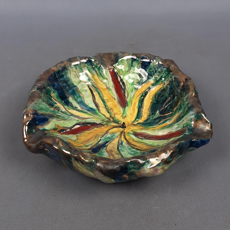 Puede incluir: Un cuenco de cer&aacute;mica pintado a mano con una forma org&aacute;nica &uacute;nica. El cuenco presenta una vibrante paleta de colores verde, amarillo, rojo y azul, con un borde marr&oacute;n oscuro. El dise&ntilde;o recuerda a una flor o una hoja estilizada.