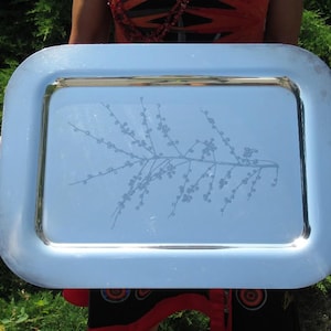 Op de afbeelding: Een rechthoekig dienblad van zilverkleurig metaal met afgeronde hoeken en een gepolijst, reflecterend oppervlak. Het midden van het dienblad is voorzien van een fijne, geëtste lijntekening van een tak met kleine besjes of knoppen.
