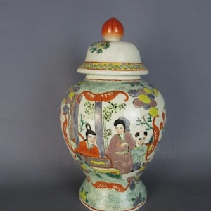 Op de afbeelding: Een keramische gemberpot met een deksel voorzien van een perzikvormige knop. De pot is versierd met een handgeschilderde scène van drie figuren in traditionele kleding, omringd door gestileerde bomen, gebladerte en bloemmotieven in oranje, groen en geel.