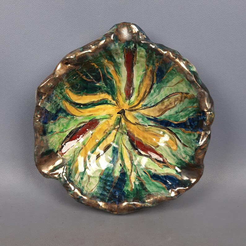 Puede incluir: Un plato de cer&aacute;mica pintado a mano con una forma org&aacute;nica &uacute;nica. El interior presenta un dise&ntilde;o vibrante con tonos amarillos, verdes, rojos y azules. El borde est&aacute; acentuado con un acabado dorado met&aacute;lico, agregando un toque de elegancia a esta pieza decorativa.
