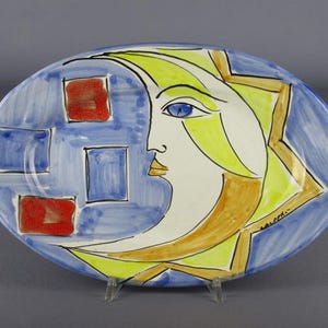 Pode incluir: Uma travessa oval de cerâmica apresentando o perfil estilizado de um rosto de lua crescente em amarelo e branco. O fundo é azul com três quadrados azuis e dois vermelhos. O design inclui contornos castanhos e a assinatura "LALOU" em preto.
