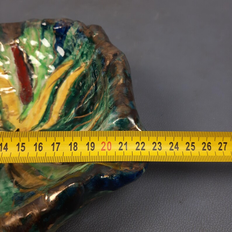 Puede incluir: Un plato decorativo de cer&aacute;mica colorida con una superficie texturizada. Presenta una mezcla de tonos verdes, amarillos, rojos y azules. Una cinta m&eacute;trica amarilla se coloca sobre el plato, mostrando una longitud de aproximadamente 35,5 a 68,5 cm.