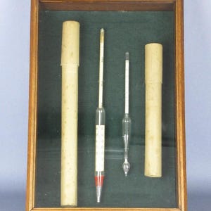May include: A framed display featuring two glass hydrometers and two cylindrical paper tubes. The hydrometers have white and red markings. The frame is wooden with a dark green background.
