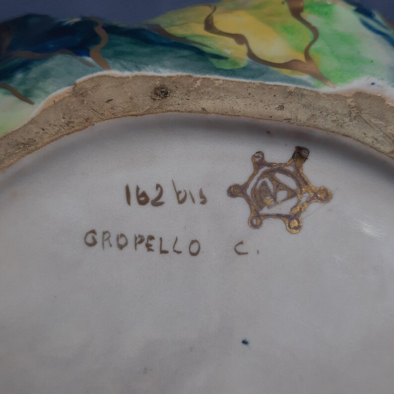 Puede incluir: Primer plano del interior de un cuenco de cer&aacute;mica con dise&ntilde;os florales pintados en azul, verde y amarillo. El borde del cuenco tiene una textura rugosa y sin esmaltar. La base es blanca con el texto "162 bis GROPELLO C." y un emblema dorado.