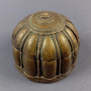 May include: A bronze-coloured, dome-shaped object with intricate detailing. The top features a floral design surrounded by a circular pattern. Vertical ridges and decorative lines adorn the sides, creating a textured appearance. The object appears to be made of metal.