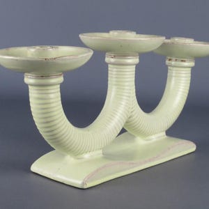 Peut inclure: Un candélabre en céramique vert pâle doté de trois bougeoirs circulaires soutenus par des bras nervurés en forme de U. La structure repose sur une base rectangulaire avec une finition lisse et des détails bruns subtils sur les bords.