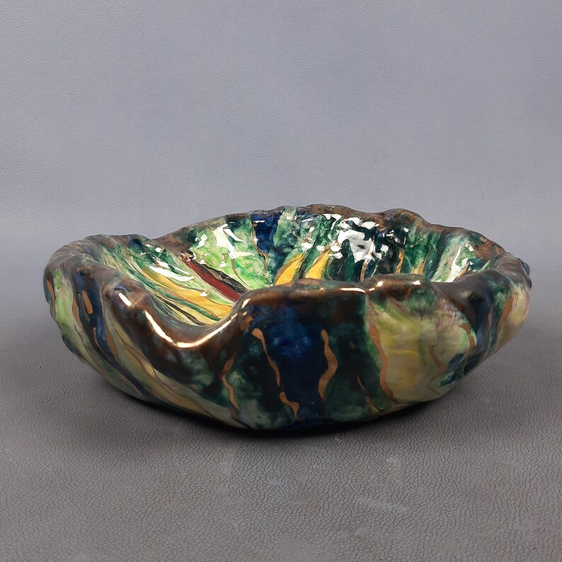 Puede incluir: Un cuenco de cer&aacute;mica hecho a mano con una forma org&aacute;nica &uacute;nica. El cuenco presenta una vibrante paleta de colores verde, azul, amarillo y marr&oacute;n, con un acabado brillante. El borde tiene un borde texturizado e irregular, lo que aumenta su atractivo art&iacute;stico.