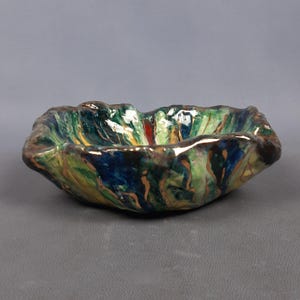 Puede incluir: Un cuenco de cer&aacute;mica hecho a mano con una forma org&aacute;nica &uacute;nica. El cuenco presenta una vibrante paleta de colores verdes, azules y dorados, con un acabado brillante. El borde tiene un borde texturizado e irregular, lo que aumenta su atractivo art&iacute;stico.
