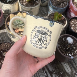May include: A white ceramic mug with a black and white graphic of a bat wing and the text "Bat Wing Tonic Elixir".