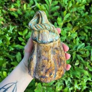 May include: A ceramic gourd with a green and brown glaze. The gourd has a unique, twisted top and a textured surface.