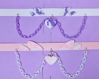 May include: Two delicate chokers with purple and white chains and heart pendants. The pendants are made of white and purple clay with a marbled effect. The chokers are adorned with white and pink butterfly wings.