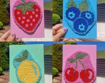 May include: Four small, colorful canvas artworks featuring embroidered fruit designs. The artworks include a red strawberry on pink, blueberries on blue, a yellow lemon on green, and red cherries on pink. The embroidery is textured and detailed.