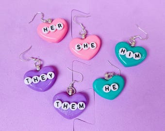 May include: Six pairs of heart-shaped earrings with different pronouns spelled out in white letters. The earrings are pink, purple, and teal.