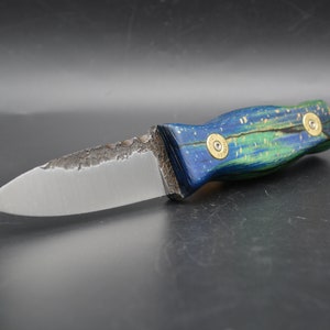 Puede incluir: Un cuchillo hecho a mano con un mango de madera azul y verde y una hoja plateada. El mango está decorado con dos casquillos de bala de latón.