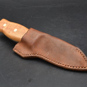 May include: A brown leather sheath with a stitched edge, designed to hold a knife with a wooden handle. The handle has two white dots.