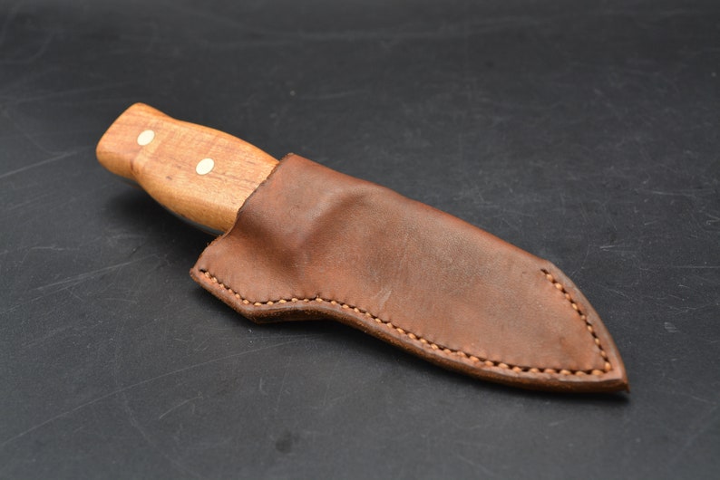 May include: A brown leather sheath with a stitched edge, designed to hold a knife with a wooden handle. The handle has two white dots.