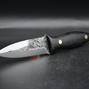 Peut inclure: Un couteau à lame fixe doté d'une lame en acier pointue avec une finition martelée sombre et texturée près de la garde et un tranchant lisse et poli. Le manche est en bois sombre et fixé par deux goupilles en laiton circulaires.