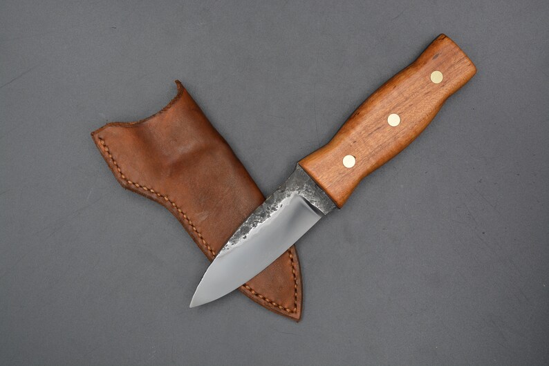 May include: A small, handmade knife with a brown wooden handle and a leather sheath. The blade is made of steel and has a slight curve.