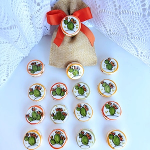 May include: A collection of decorative ceramic buttons featuring a cactus design. The buttons have a white base with a green cactus and orange accents. A burlap bag with an orange ribbon and a matching button is also present.