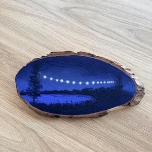 Può includere: Scena di paesaggio dipinta a mano su una fetta di legno. L'opera raffigura una scena notturna del lago con una fila di luci. La fetta di legno ha un bordo di corteccia naturale ed è ambientata su uno sfondo di venature del legno chiaro.