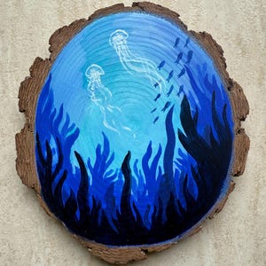 Può includere: Fetta di legno dipinta a mano con una scena sottomarina. Due meduse fluttuano in un oceano blu e turchese, circondate da alghe blu scuro e un banco di pesci. Il bordo naturale della corteccia del legno incornicia l'opera d'arte.