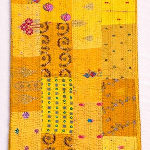 May include: A rectangular, patchwork textile in shades of yellow, with intricate embroidered floral and geometric patterns. The design features a variety of textures and colors, including pink, blue, and green accents. The piece is likely a decorative quilt or throw.