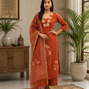 May include: A rust-colored three-piece outfit consisting of a straight-cut kurta with three-quarter sleeves, wide-leg trousers, and a matching sheer dupatta. The ensemble features cream-colored floral block print motifs and gold trim.