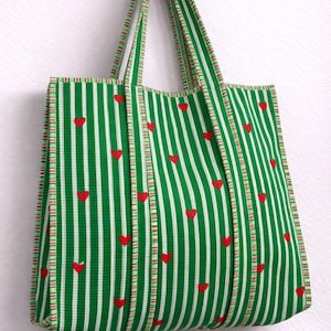 Puede incluir: Bolso tote a rayas verdes y blancas con detalles de corazones rojos. El bolso presenta rayas verticales y formas de corazón rojo dispersas por la tela. Las asas y los bordes están ribeteados con un patrón de rayas a juego.