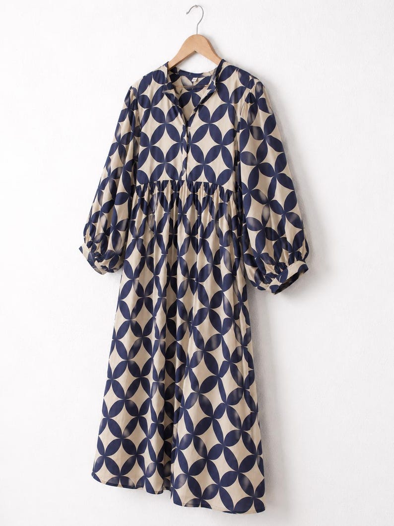 May include: A midi-length dress featuring a beige base with a repeating dark navy blue circular geometric pattern. The dress has long, loose sleeves with gathered cuffs, a gathered waistline, and a simple collarless neckline with a front button placket.
