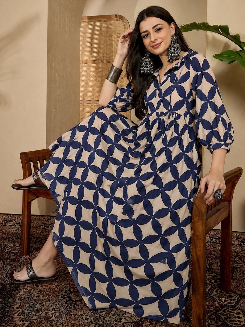 May include: A long-sleeved maxi dress featuring a repeating navy blue geometric circle pattern on a beige background. The dress has a collared neckline, a gathered waistline, and voluminous sleeves that extend to the elbows, creating a flowing silhouette.