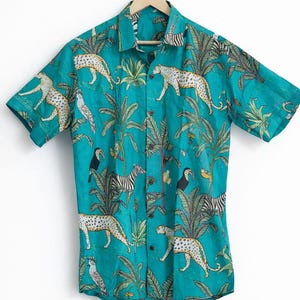 Puede incluir: Camisa turquesa con estampado de jungla. La camisa de manga corta presenta leopardos, cebras, tucanes y palmeras. La camisa está colgada de una percha de madera.