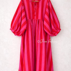 May include: A knee-length dress featuring vertical stripes in alternating shades of bright red and hot pink. The garment has voluminous puff sleeves, a round neckline with a small tie closure at the centre, and a gathered waistline. The text Shree Fashion is visible.