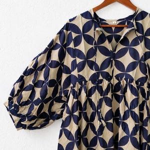 May include: A beige dress featuring a repeating dark navy blue circular geometric pattern. The garment has a V-neckline with thin tie closures, voluminous long sleeves with gathered cuffs, and a gathered waistline, all displayed on a wooden hanger.