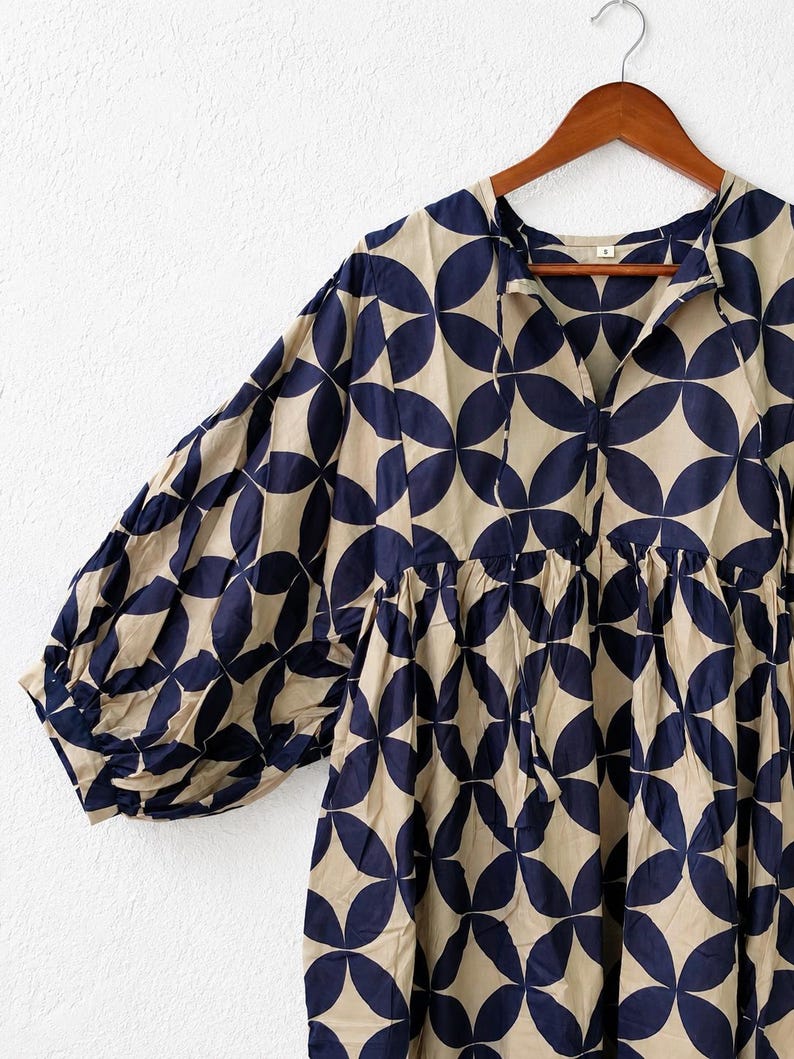 May include: A beige dress featuring a repeating dark navy blue circular geometric pattern. The garment has a V-neckline with thin tie closures, voluminous long sleeves with gathered cuffs, and a gathered waistline, all displayed on a wooden hanger.