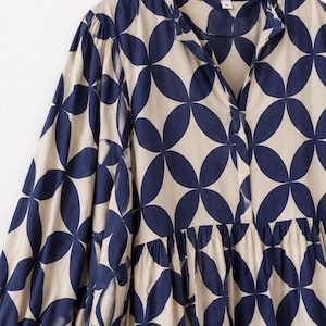 May include: A long-sleeved blouse featuring a repeating navy blue geometric petal pattern on a beige background. The garment has a split V-neckline, a gathered waist seam, and full sleeves with cuffs. A small white tag with the letter M is visible.
