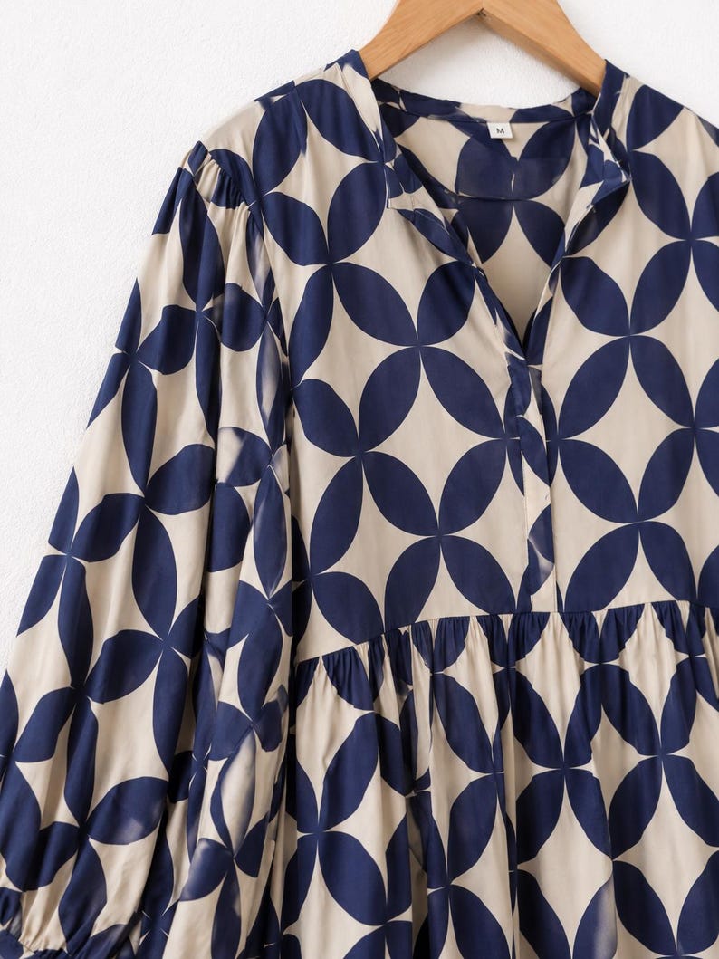 May include: A long-sleeved blouse featuring a repeating navy blue geometric petal pattern on a beige background. The garment has a split V-neckline, a gathered waist seam, and full sleeves with cuffs. A small white tag with the letter M is visible.