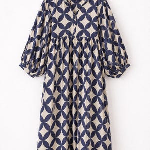 May include: A midi-length dress featuring a beige base with a repeating dark blue geometric petal pattern. The dress has a split V-neckline, a gathered waistline, and long, voluminous sleeves with elasticized cuffs, all displayed on a wooden hanger.